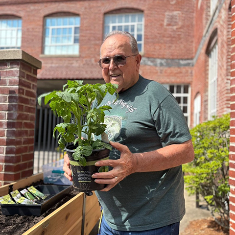 An older gentleman smiling and wearing sunglasses and holding a potted plant outside a brick building.