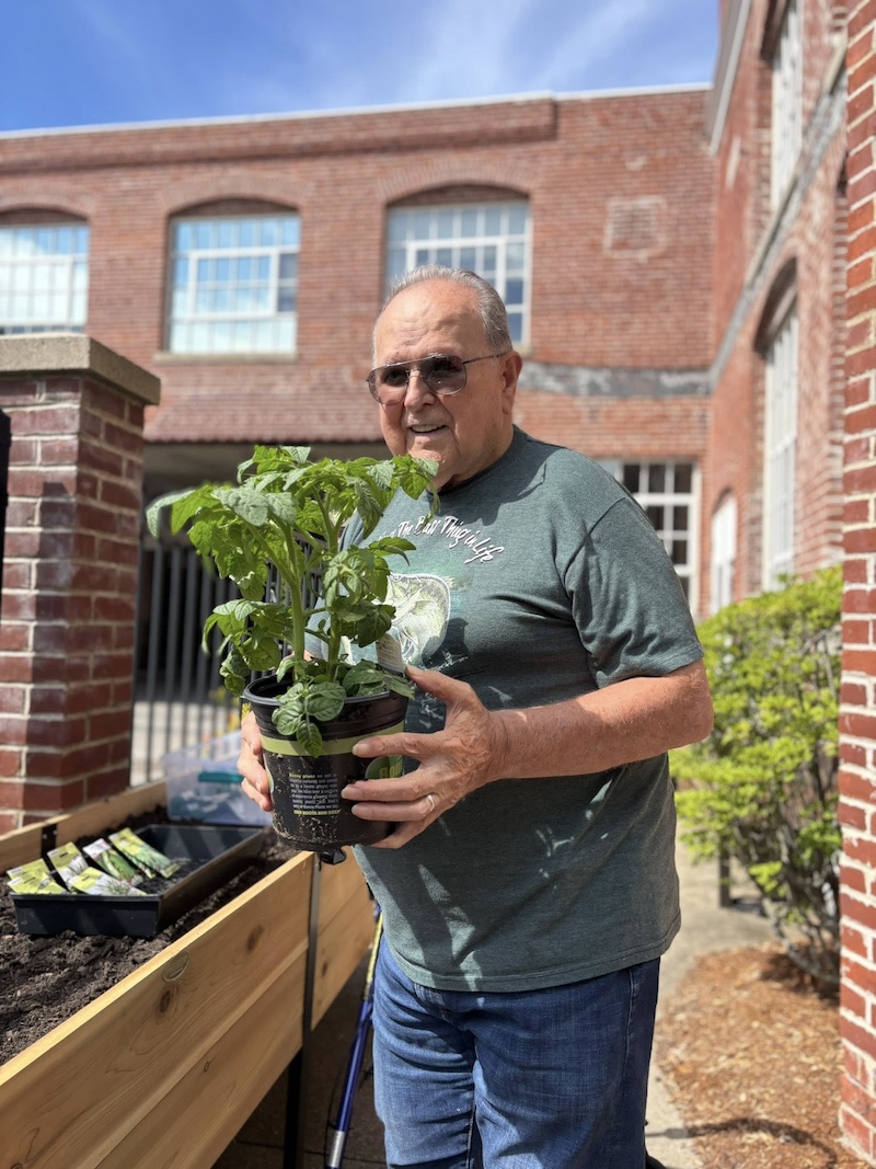 Benefits of Respite Care An older gentleman smiling and wearing sunglasses and holding a potted plant outside a brick building.
